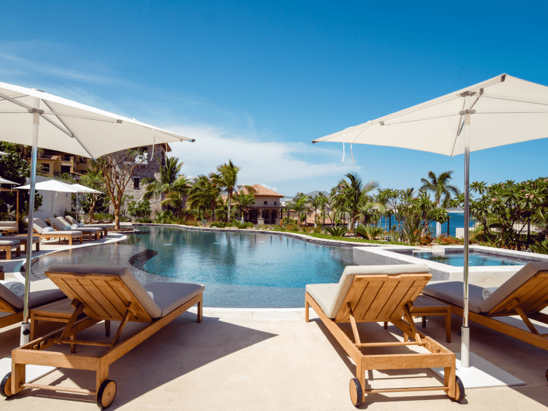 Reclined lounge chairs with white square shaped umbrellas overhead for shade, face inwards toward the pool