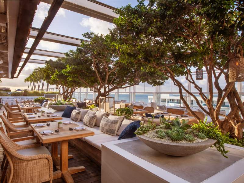 An outdoor dining area with wooden tables and chairs, surrounded by lush greenery and decorative cushions also the space offers a view of the ocean and is partially shaded by a pergola