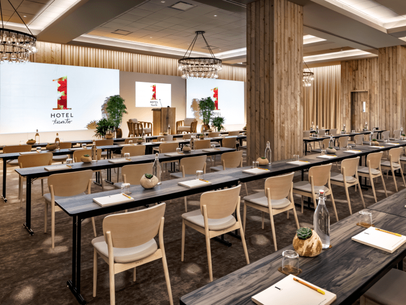 Black wooden tables stretch lengthwise across a 1 Hotels meeting room.  Three projector screens line the wall in which all seats are facing.