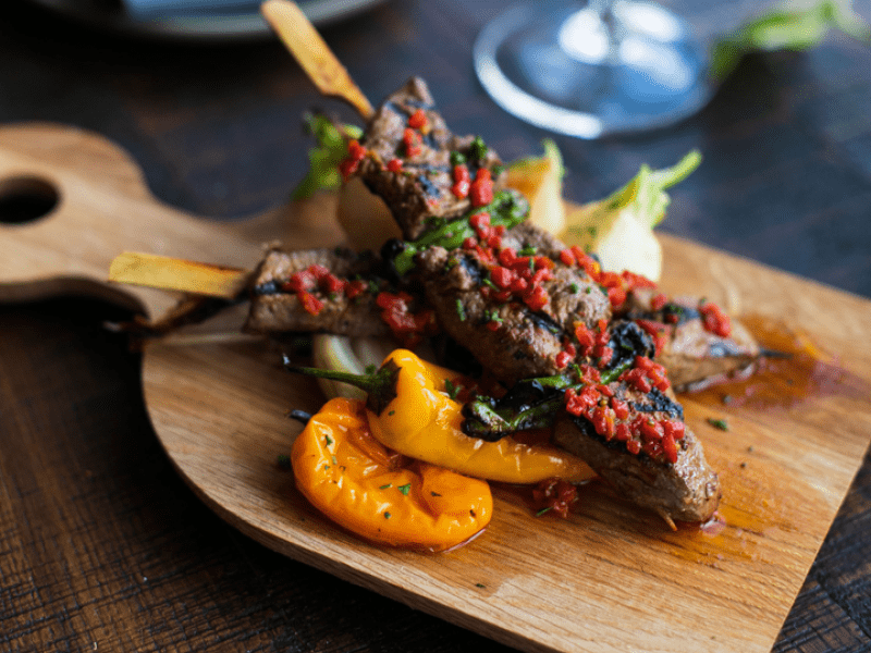Two grilled meat skewers complimented by a side of roasted peppers and a glass of red wine