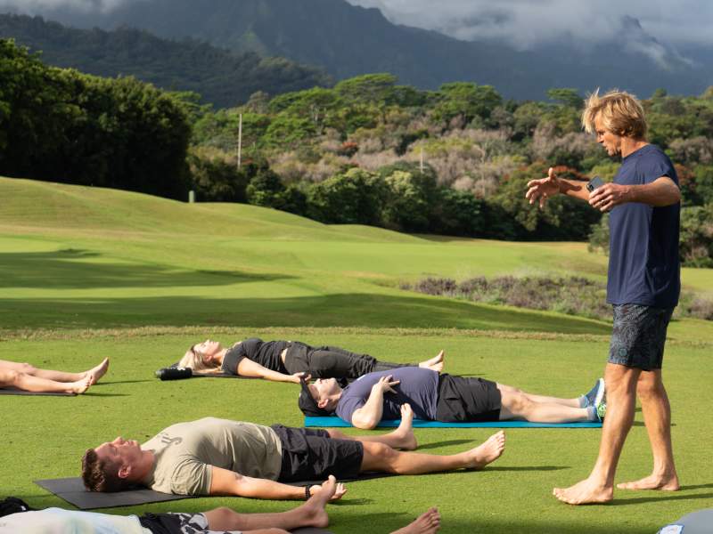 Group of people laying on yoga mats outside while an instructor guides them