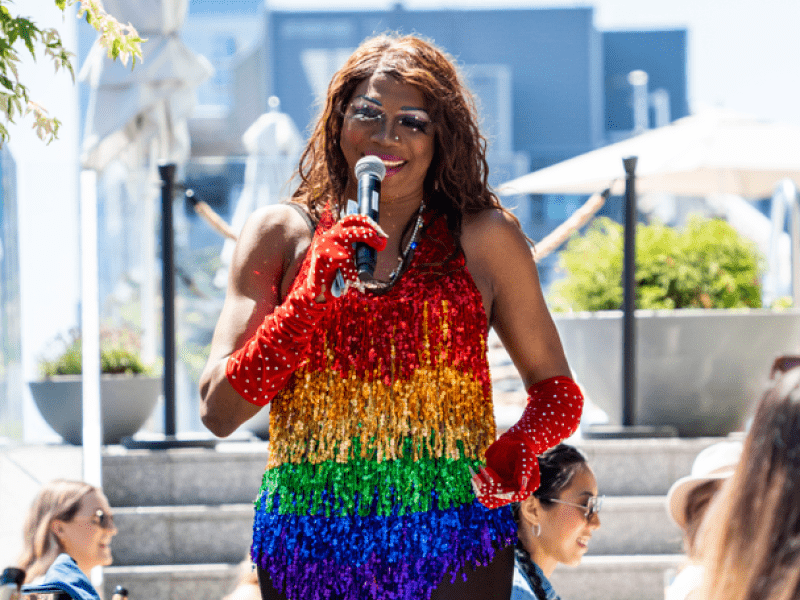 Person wearing a rainbow bead dress with red gloves speaking into a microphone