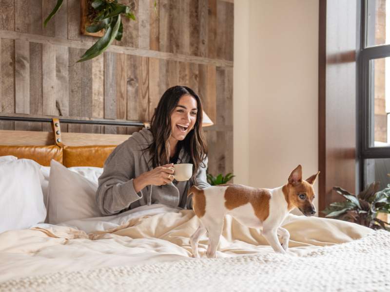 A woman holding a coffee cup smiles in bed while her small dog plays on the bed.