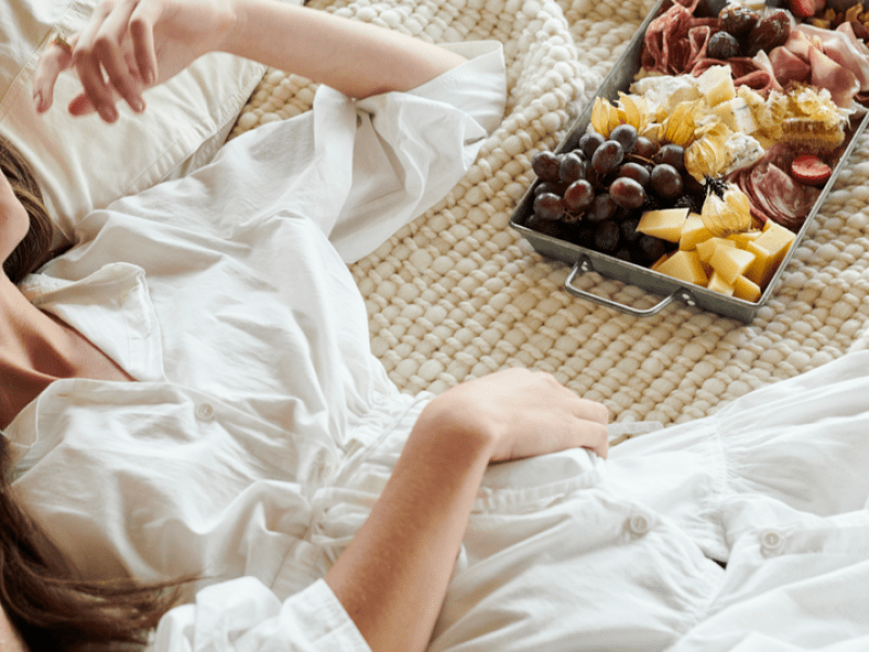 A woman in a white robe lounges on her bed next to a charcuterie plate