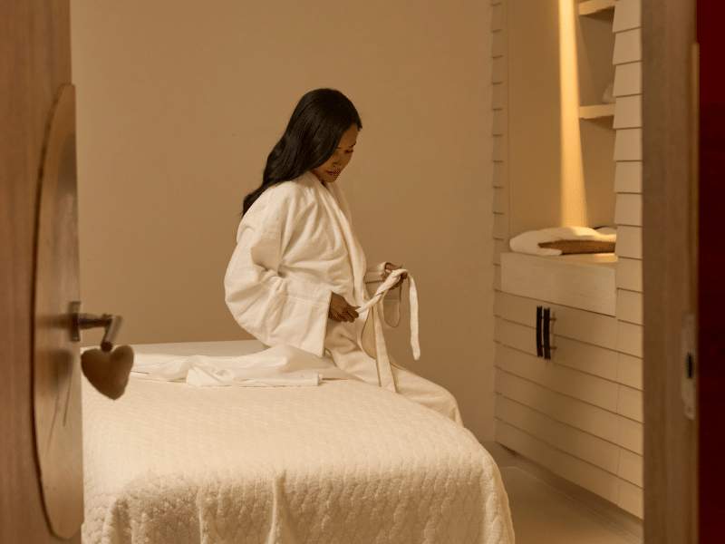 Bamford Wellness Spa treatment room with a woman in a white robe on the edge of a bed, shelving behind
