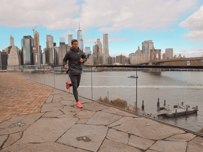 A man in black athleisure runs along the waterfront.  Views of the city skyline are visible behind him.