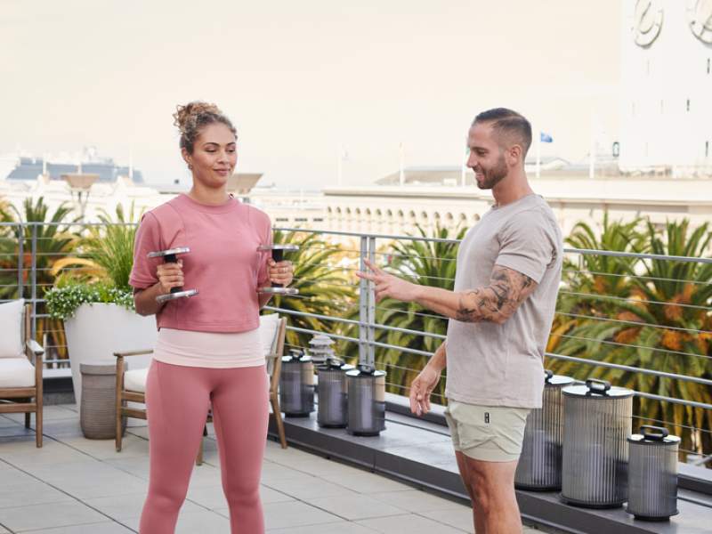 Un entraîneur privé donne des instructions à une cliente qui tient des poids libres, sur le toit du 1 Hotels SFO.