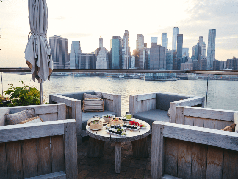 Patio with four chairs facing a table with several sushi dishes, with the harbor visible in distance