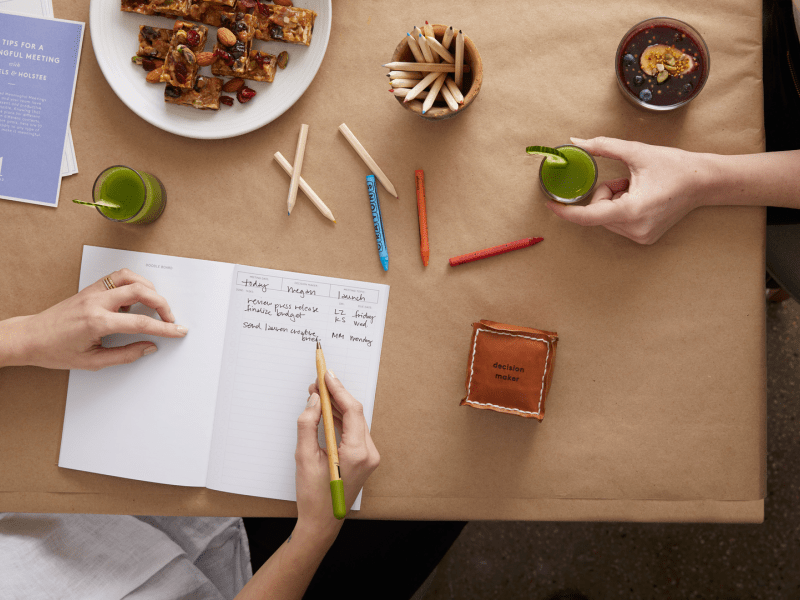 Person writing in a journal while another holds a drink