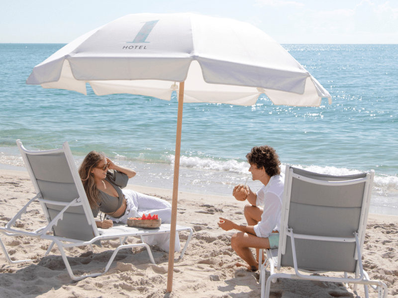 Two people sitting on beach chairs under a 1 Hotel umbrella