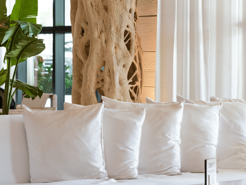 Five white pillows line an equally white sofa. A driftwood style art installation compliments the couch as a backdrop piece.