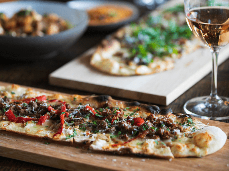 Long flatbread pizzas served on wooden cutting boards, paired with a glass of white wine