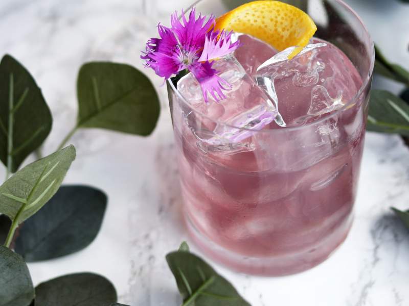A pink cocktail with a lemon wedge and flower garnish