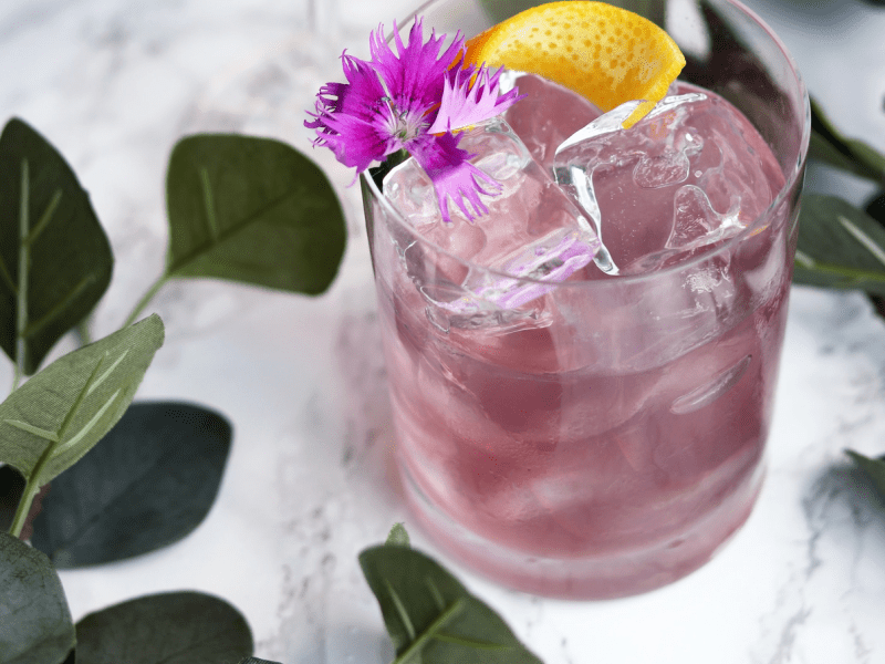 A pink cocktail with a lemon wedge and flower garnish