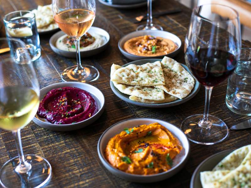 A dark wooden table set with a spread of various dips, pita, and wine