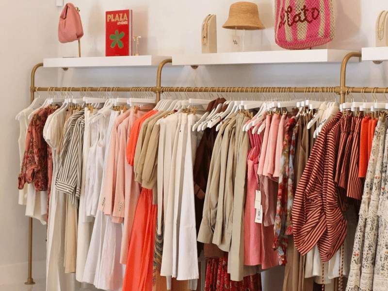 Clothes on a rack in a boutique with bags and hats on shelf above
