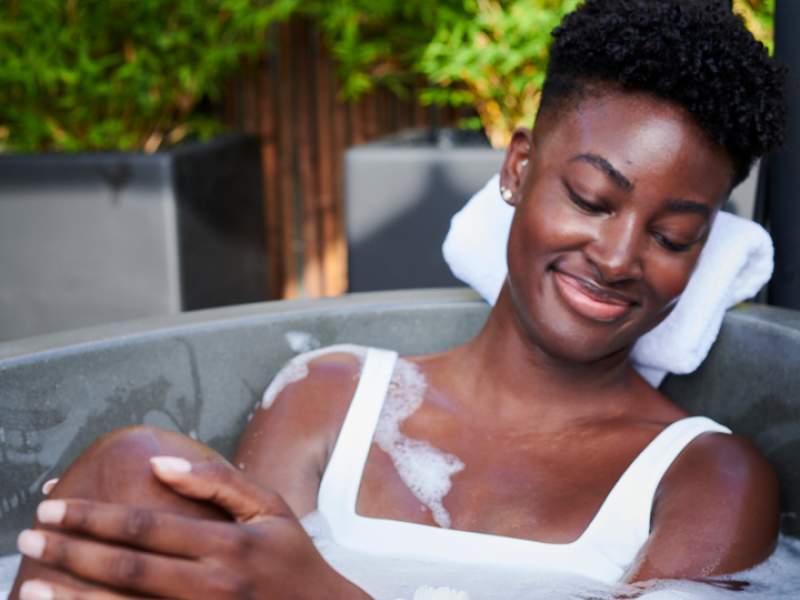 A woman in a white one piece bathing suit relaxes in an outdoor bathtub.