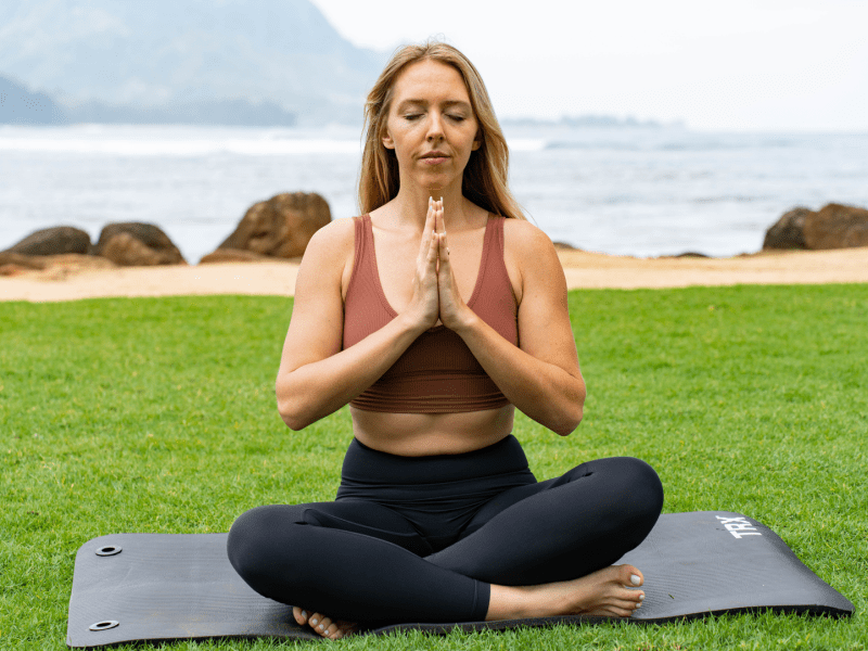 Person sitting on a yoga mat in a field with their hands together