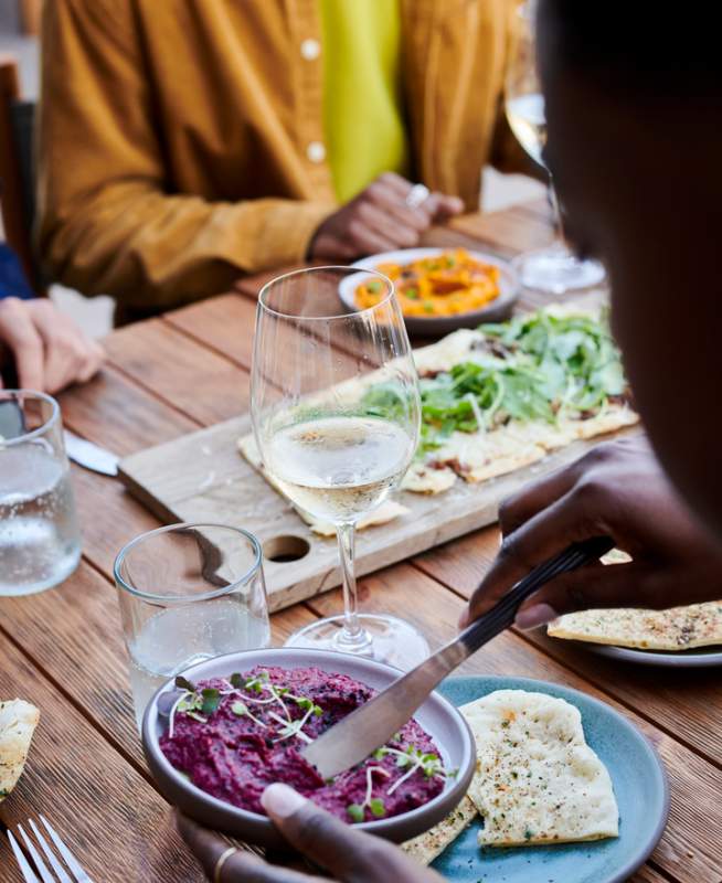 People seated around a wooden table are sampling a variety of dips with pita bread