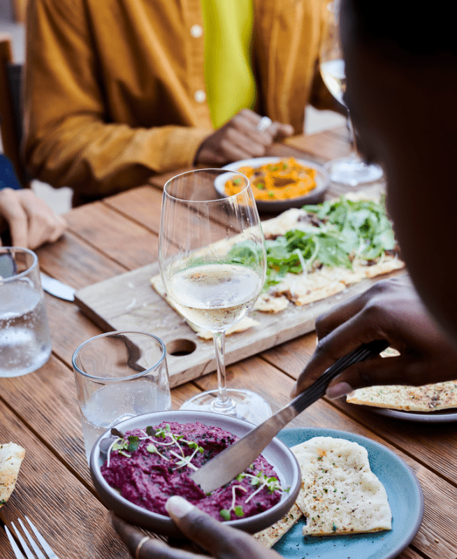 People seated around a wooden table are sampling a variety of dips with pita bread
