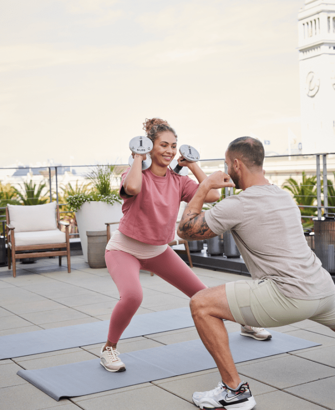 A male private trainer instructs a female client holding free weights, performing a side squat