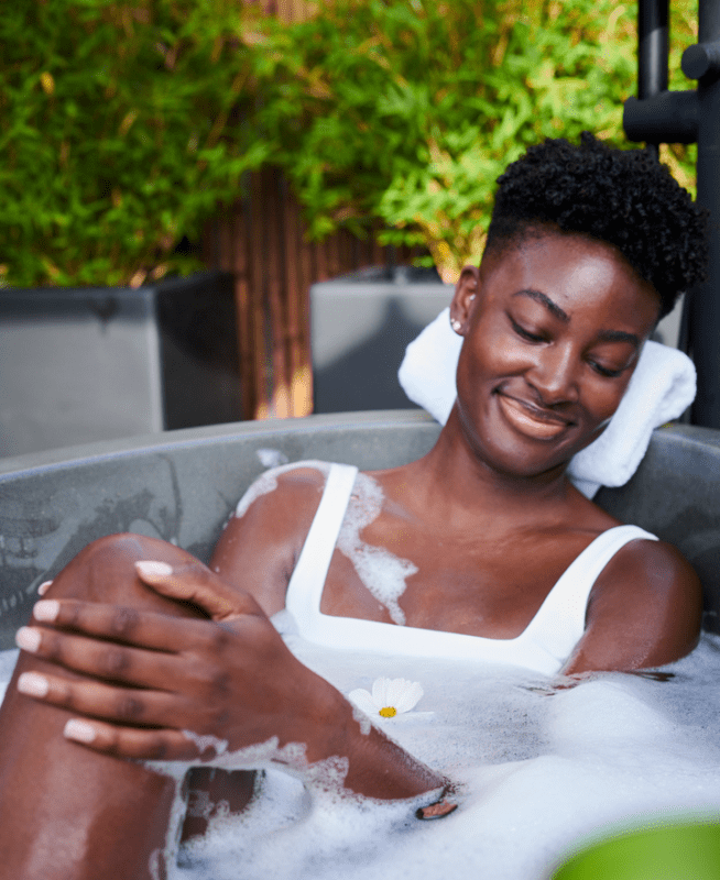 A woman in a white one piece bathing suit relaxes in an outdoor bathtub.