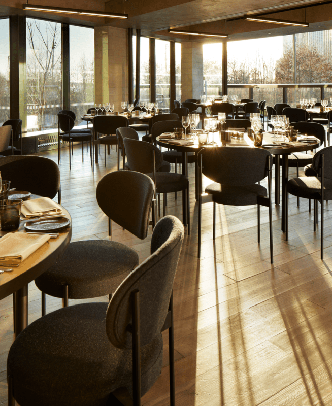 Aster Social Evening.  A spacious room with large round wooden tables each set for eight individuals.  Sunlight pours in through the windows.