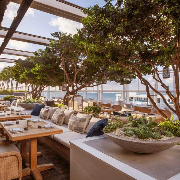 무성한 녹지와 장식용 쿠션으로 둘러싸인 나무 테이블과 의자가 있는 야외 식사 공간은 바다 전망을 제공하며 퍼걸러로 부분적으로 그늘이 드리워져 있습니다.