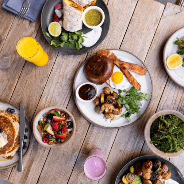 A full table spread of various breakfast dishes, including from left to right, a stack of blueberry pancakes, a breakfast wrap, bacon and eggs, and a salad.