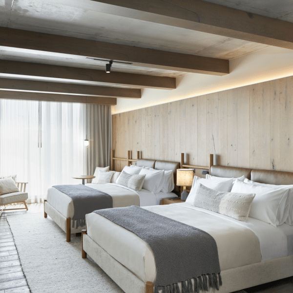 Two queen size beds with wooden headboards sit side by side basking in the sun shining through the nearby window