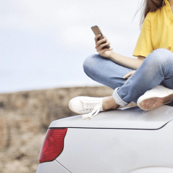 A woman wearing a yellow polo and faded blue jeans sits cross legged atop a white car with its top dropped back
