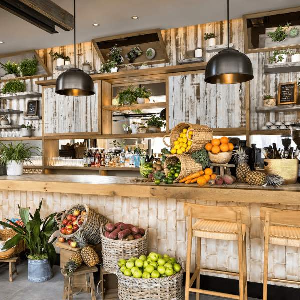 Baskets of fruit decorate a lengthwise wooden bar with stool seating.  Displays of drinks, coffee machines, dishes, and food can be seen lining the wall behind the bar.