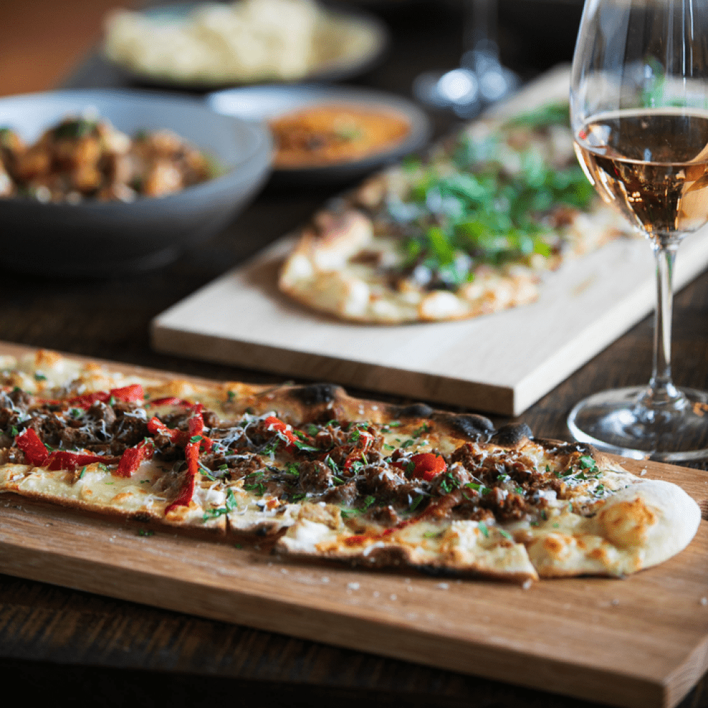 Long flatbread pizzas served on wooden cutting boards, paired with a glass of white wine
