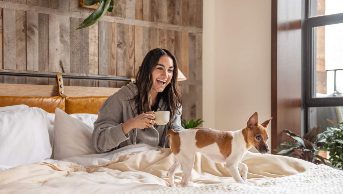 A woman holding a coffee cup smiles in bed while her small dog plays on the bed.
