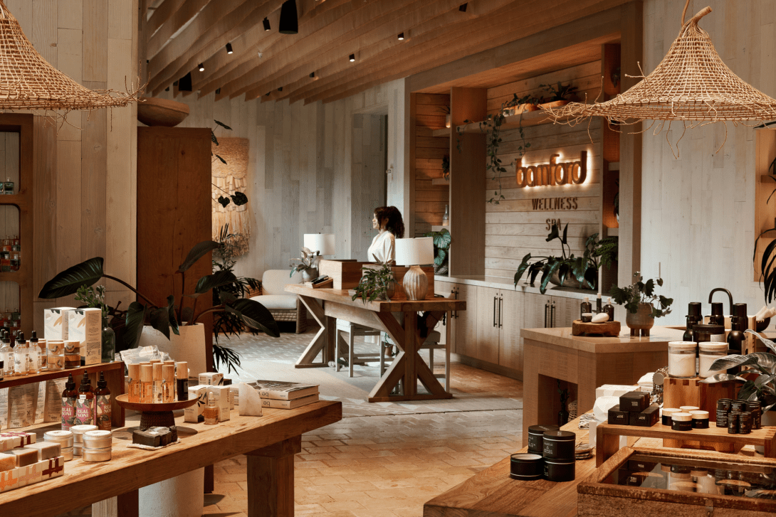The Bamford Wellness Spa entrance room, timber tables of skincare products under woven pendant shades, with the lit spa name on the pale wood wall behind.