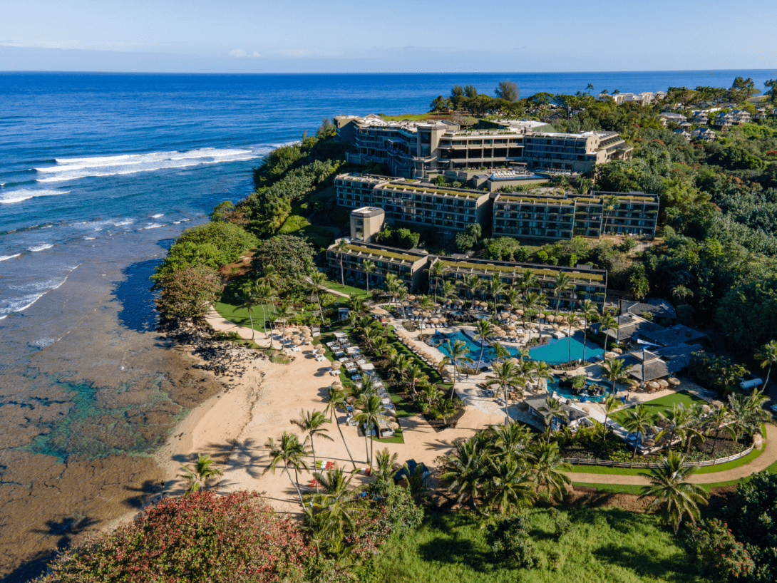 An aerial view of the resort stepping down a green headland above Hanalei Bay, its pools and palms set between the buildings and a reef-fringed beach.