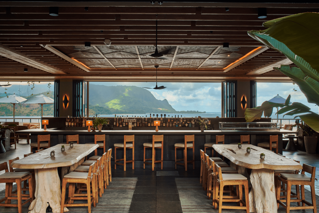 The sushi counter at Welina, a long timber bar with woven-back stools and live-edge tables, its whole far wall open to the mountains across the bay.