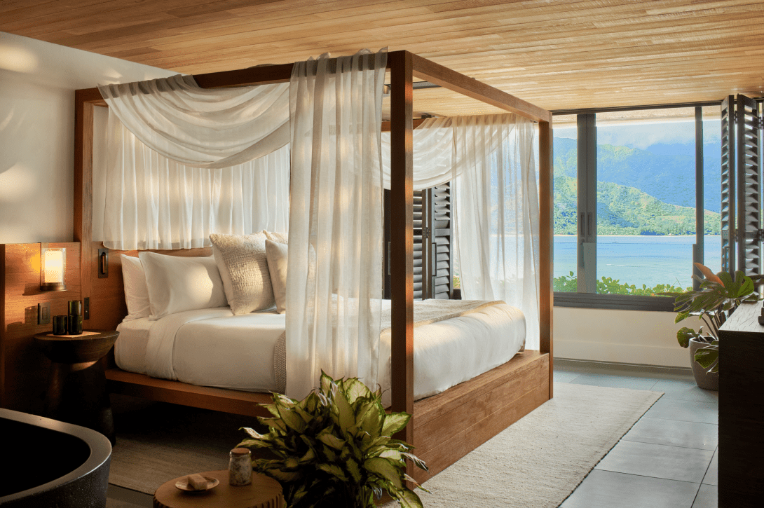 An ocean loft with a timber four-poster bed draped in white gauze under a slatted wood ceiling, its sliding doors open to the bay and green mountains.