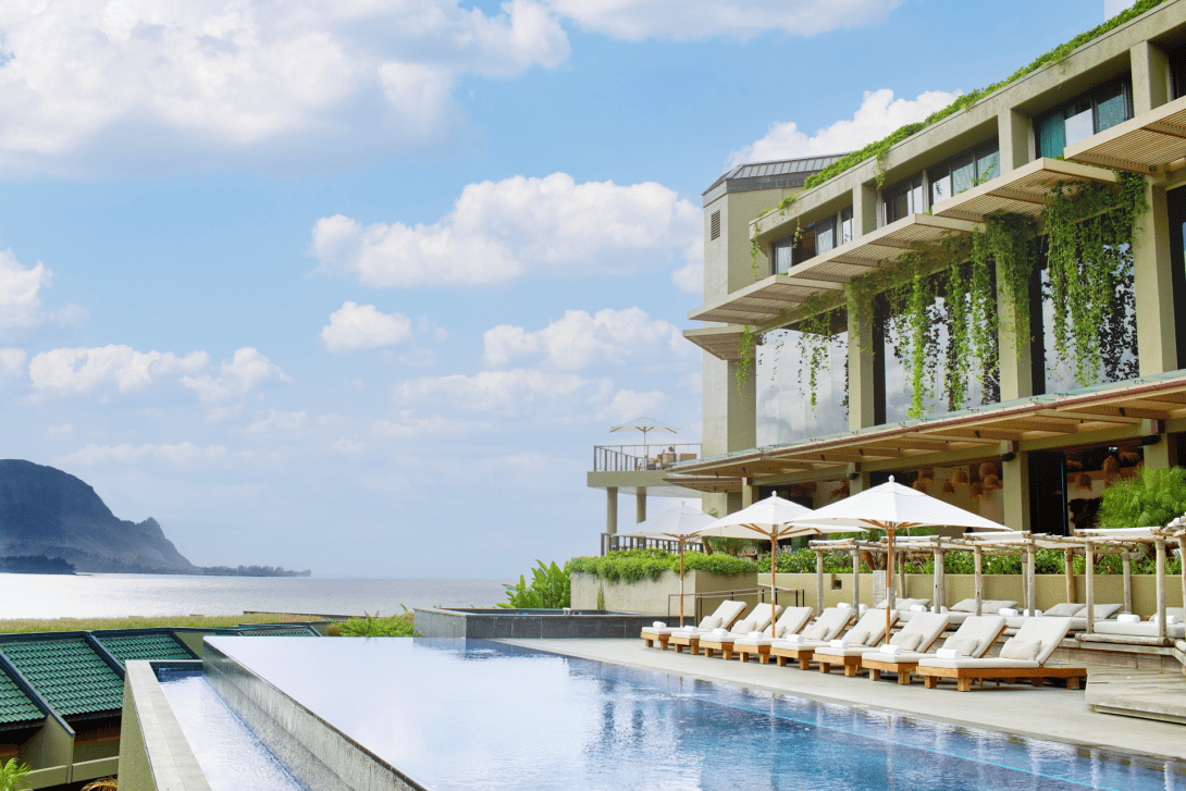 The adult infinity pool on a terrace above the ocean, a row of white loungers and parasols beside it and trailing greenery hanging from the balconies above.