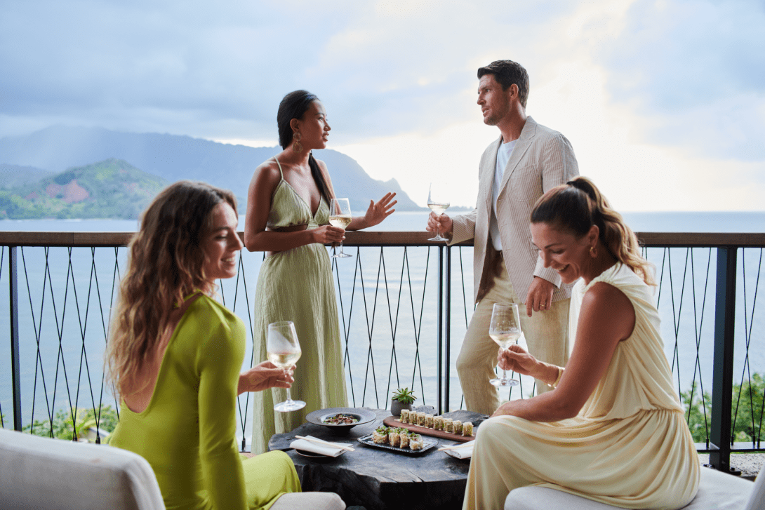 Four guests with wine around a low table of sushi on the Welina terrace, the bay and its green ridges behind the railing.