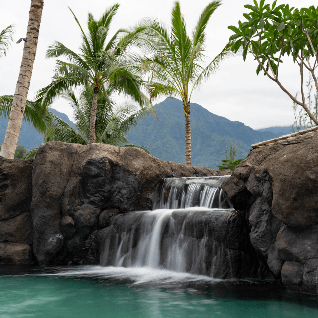 Water falling in two sheets over dark volcanic boulders into the green main pool, with palms and a ridgeline rising behind.