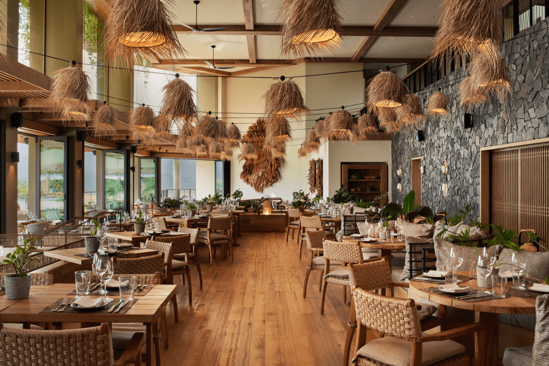 1 Kitchen laid for service, timber tables and woven chairs on a wood floor beneath a ceiling hung with dozens of shaggy fibre pendants, beside a rough stone wall.