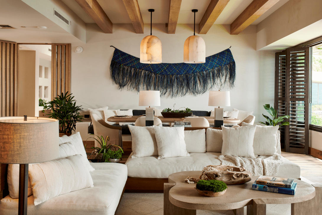 The penthouse living room under exposed timber beams, deep cream sofas around organic-shaped low tables, with a fringed indigo textile hung between two alabaster pendants.