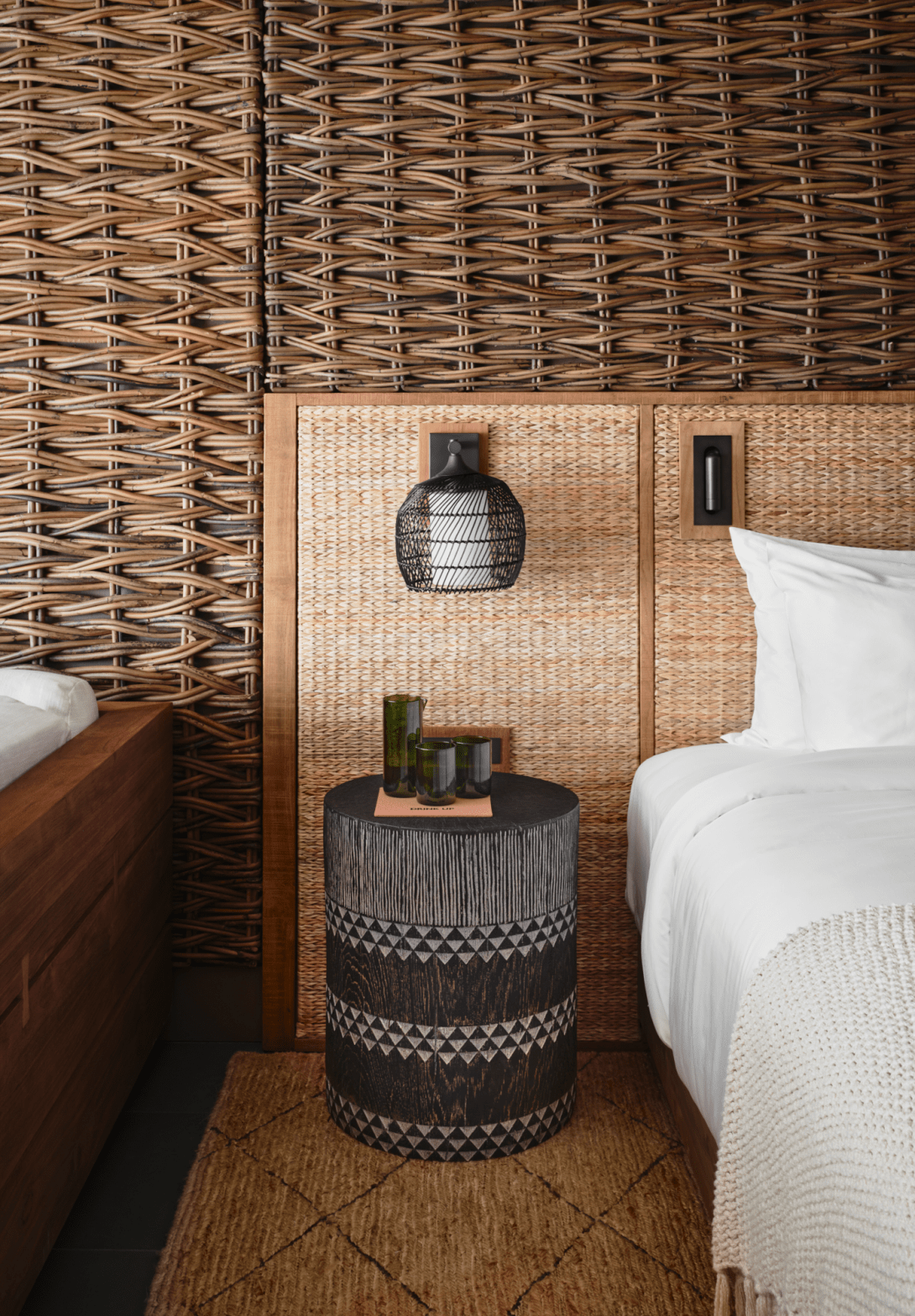 A bedside detail: a drum table of dark woven fibre beside a white-dressed bed, against a headboard wall of rattan panels and coarse basketry.