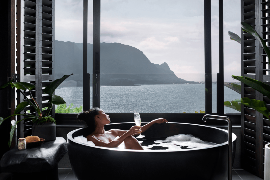 A guest in a round black soaking tub raising a glass, framed by louvred shutters that open onto the bay and the Napali ridgeline.