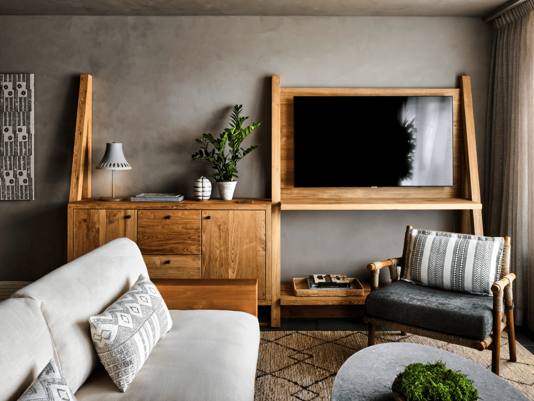 A room's sitting area, a cream sofa and grey armchair on a woven rug facing a timber console and a screen mounted on a ladder-style frame.