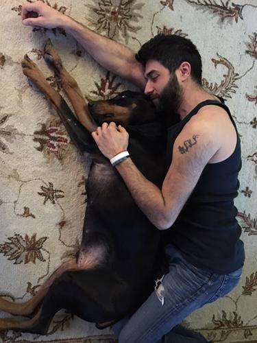 A man in jeans and a tank top lying on a bed spooing his doberman pinscher