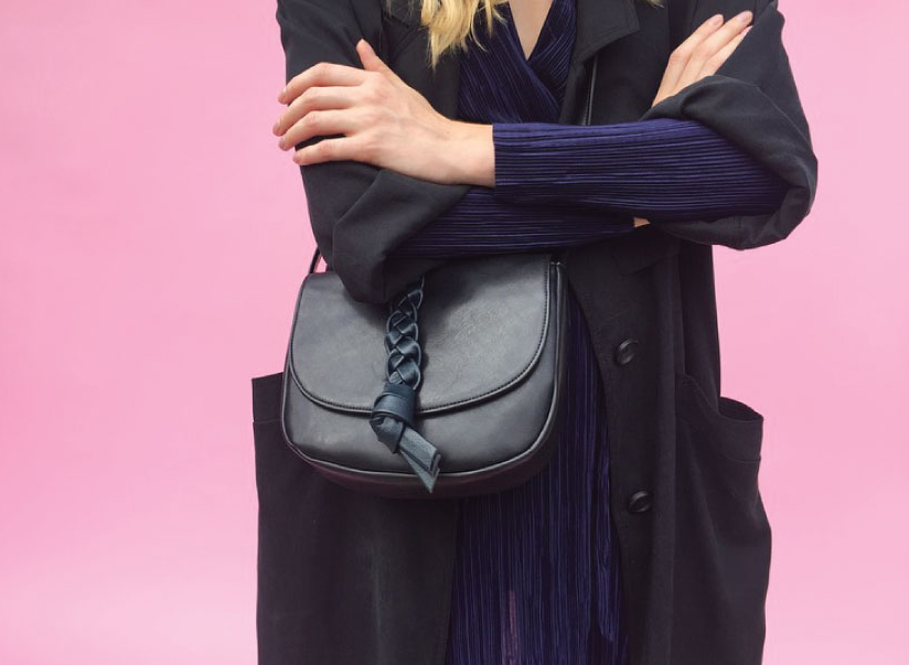 A woman in a long black coat and a black leather bag standing in front of a pink wall