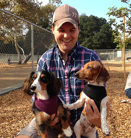 A man in a park holding two pet daschunds and smiling