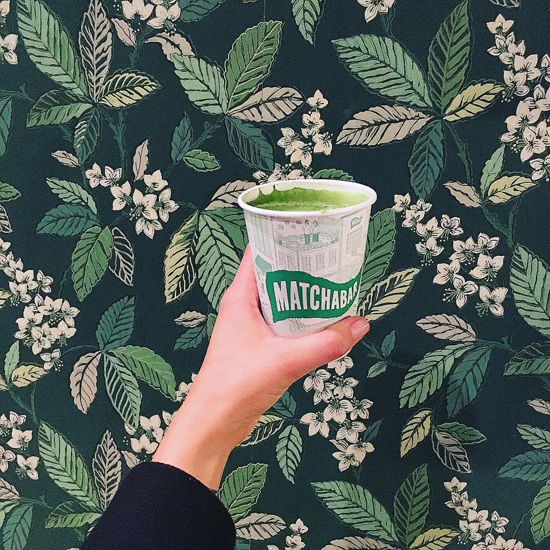 A green matcha tea in a Matcha Bar cup held up in front of leafy green wallpaper